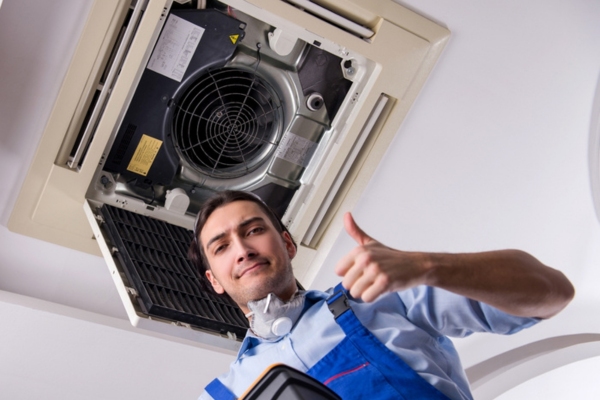 HVAC technician doing a thumbs up below a ceiling air conditioner depicting the importance of air conditioner maintenance